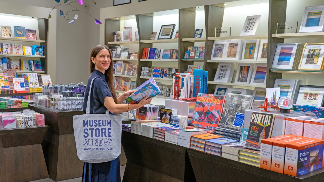 A person browsing in the book section of a museum store while holding a tote bag reading "Museum Store Sunday: Shop With Purpose"