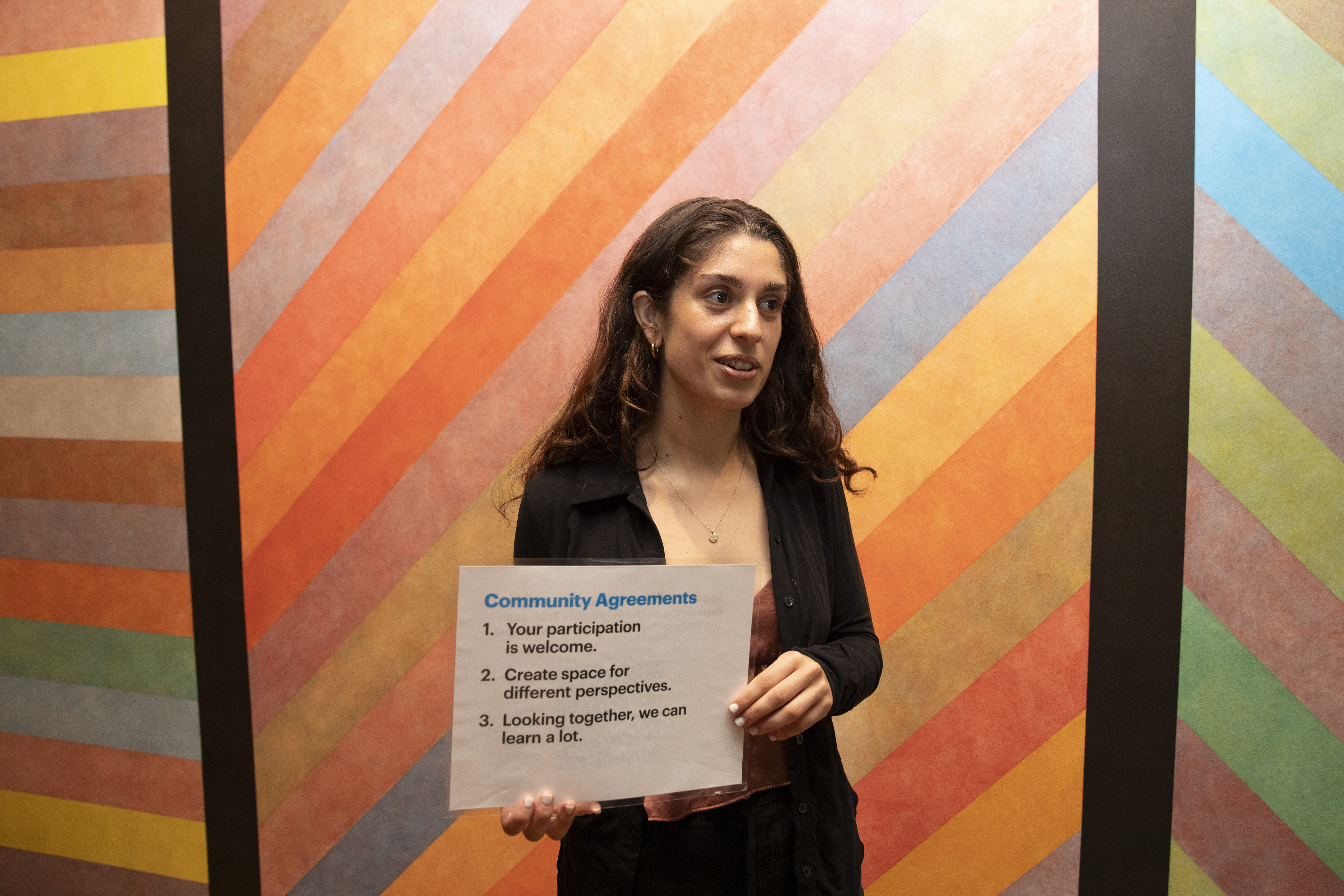 A person in front of an artwork holding a sign that reads "Community Agreements: 1. Your participation is welcome. 2. Create space for different perspectives. 3. Looking together, we can learn a lot."